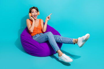 Full size photo of crazy astonished girl sit on bean bag touch headphones directing at sale empty space isolated on blue color background