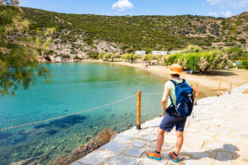 Woman tourist with sun hat and backpack on coastal path to Marina Gialos beach, Sifnos island,...