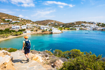 Woman tourist with sun hat and backpack walking on coastal path to Marina Gialos beach, Sifnos...
