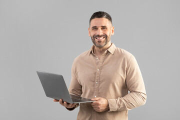 A man is holding a laptop computer in his hands, looking at the screen with a smile on his face. He...