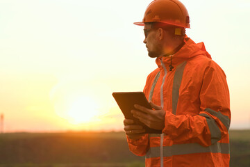 An engineer in a construction helmet, goggles and raincoat, stands with a tablet in his hands...