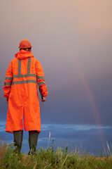 A man in a raincoat, a protective helmet and boots against a background of cloudy sky and a rainbow