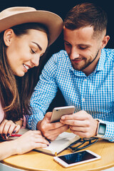 Smiling male and female best friends making booking together on web page using modern mobile phone in cafe,couple in love watching funny videos online in networks via telephone having fun on date