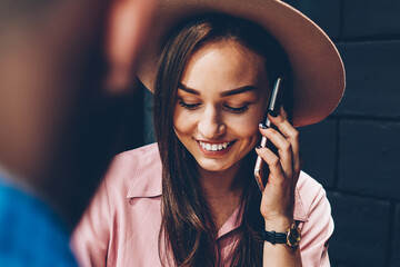 Cheerful trendy dressed woman talking on cellular during meeting with boyfriend in cafe, hipster girl having telephone conversation about booking satisfied with communication spending time with friend