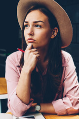 Beautiful hipster girl in trendy hat spending time in cafe interior pondering on plans for work,stylishly dressed young woman concentrated on idea for article sitting at table with pen looking away