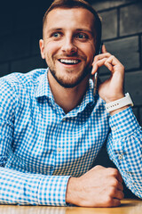 Cheerful hipster guy happy about getting good news during mobile phone conversation, successful businessman celebrating achievement of project excited while talking on telephone sitting in cafe