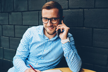 Portrait of smiling businessman noting information during phone conversation with colleague,positive male in spectacles talking with banking service operator writing in notepad looking at camera