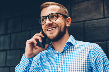 Smiling hipster guy in spectacles satisfied with tariffs for mobile phone calls spending time indoors,positive businessman exited with good news about project talking on telephone with colleague