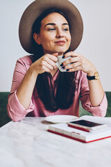 Cute hipster girl in trendy hat drinking coffee in morning enjoying starting day with favorite beverage,smiling attractive woman having work break in cafe interior thinking about plans for day