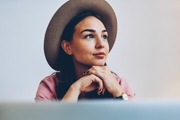Dreamy hipster girl in stylish hat enjoying free time pondering on idea for publication online, attractive young woman sitting near laptop computer thinking about designing web project indoors