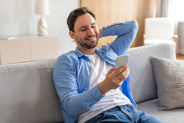 A man is sitting on a couch indoors, engrossed in using his smartphone. He is focused on the...
