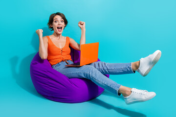 Full size photo of ecstatic girl wear orange knit top sit on bean bag with laptop scream win...