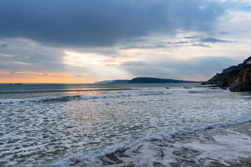 Plage de la presqu'île de Crozon, à la fin de journée, sous un ciel couvert teinté d'un rose orangé, où la mer offre ses reflets nacrés et son écume blanche, créant une scène à la fois douce et captiv