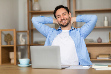 A content man leans back in his chair in a well-lit home office, his hands behind his head,...