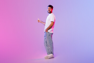 A man stands in a studio with mixed pink and blue lighting, smiling and giving a thumbs-up while...