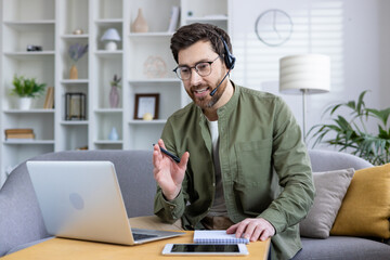 Naklejka premium Man working from home wearing a headset, holding a pen, and having a video call on his laptop while sitting on a sofa in a modern living room.