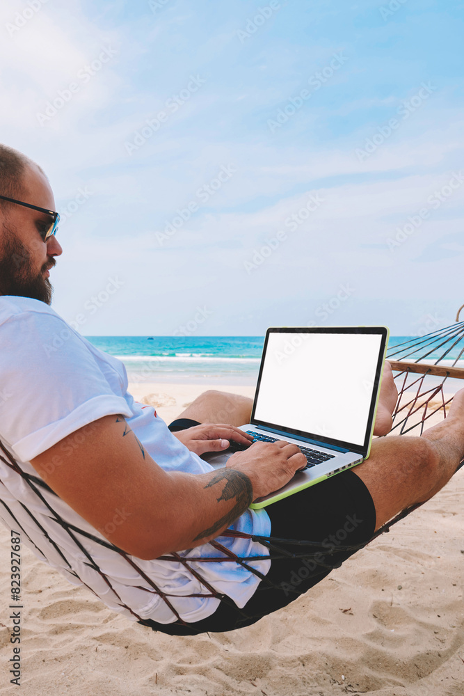Poster young bearded businessman in sunglasses rest on hammock on ocean beach while working distantly on la