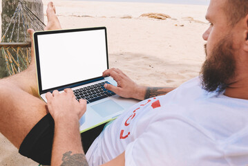 Male businessman tourist typing text on keyboard of computer with blank screen for your internet website rest on tropic island. Young man freelancer work at laptop during distance job lying on hammock