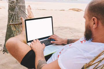 Young man resting on hammock while doing work freelance remotely at modern laptop computer with mock up blank screen for advertise content during summer vacations on tropic island with blue sea