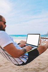 Young bearded businessman in sunglasses rest on hammock on ocean beach while working distantly on laptop computer with blank screen area for your web page advertise content during summer vacations