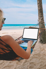 Female freelancer typing text on keyboard of netbook with blank screen area for your website during remotely work on sandy beach of tropical island.Young man chatting online on phone lying on hammock