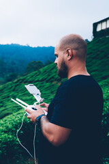 Young man controling flying dron with help remote control to shoot green landscape and natural environment of tropical island.Male bearded explower standing in tea plantation during expedition