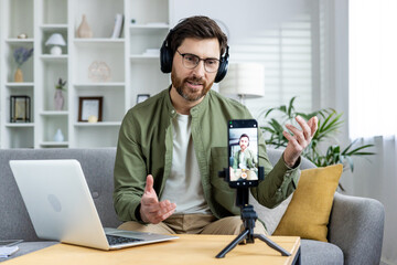 Man with headphones recording a video blog on smartphone at home, using laptop, sitting on a couch.