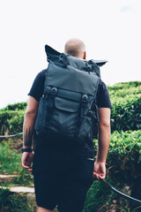Back view of young man lover of travel with black rucksack walking in tropical island with green plantation during summer trip.Male hiker with backpack during treck in asia wild environment