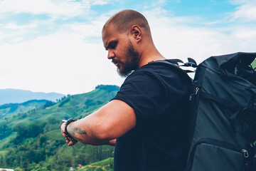 Pensive bearded tourist with backpack looking at smartwatch to tracking GPS walking in green...
