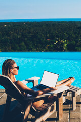 Young woman in sunglasses resting on sunbed near blue swimming pool and working freelance at modern...