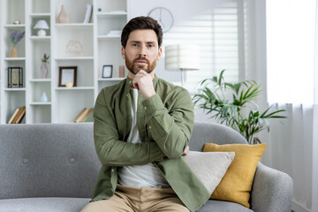 Young man in casual attire sits on a couch in a modern living room, deep in thought. Shelves and...