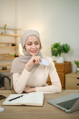 A woman wearing a scarf is sitting at a desk with a laptop and a notebook. She is smiling and holding a credit card