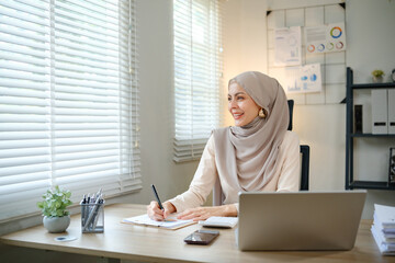 A woman wearing a head scarf is sitting at a desk with a laptop and a pen. She is smiling and she is happy
