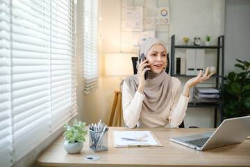 A woman wearing a hijab is talking on her cell phone in a cubicle. She is sitting at a desk with a laptop and a computer monitor