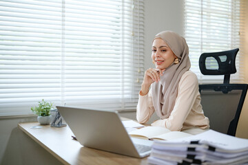 A woman wearing a hijab is sitting at a desk with a laptop and a stack of papers. She is smiling and she is focused on her work