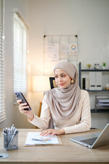A woman wearing a hijab is sitting at a desk with a laptop and a cell phone