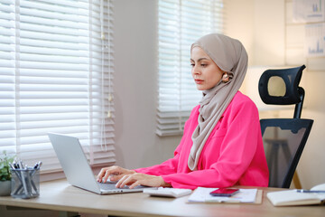 A woman wearing a pink shirt and a head scarf is typing on a laptop. She is sitting at a desk with a chair in front of her