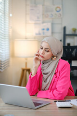 A woman wearing a pink jacket and a grey scarf is sitting at a desk with a laptop in front of her. She is focused on her work, possibly typing or browsing the internet