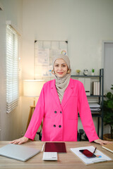 A woman in a pink jacket stands in front of a desk with a laptop, a tablet, and a stack of papers. She is a professional, possibly a businesswoman or a teacher