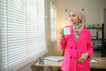 A woman in a pink jacket is holding a cup of coffee. She is standing in front of a window with a white blind