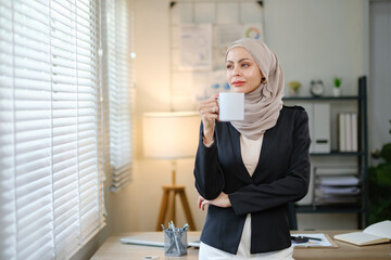 A woman in a business suit is holding a coffee cup in front of a window. She is focused and serious, possibly preparing for a meeting or working on a project