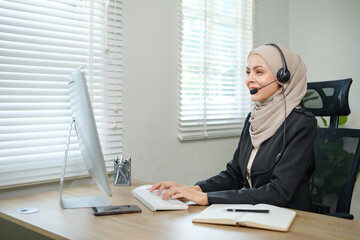A woman wearing a hijab headset is sitting at a desk in front of a computer monitor. She is typing on a keyboard and she is focused on her work
