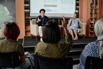 Unrecognizable female student raising hand to ask question during conference in university library,...
