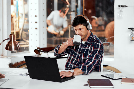 Laptop, coffee and man with headphones in workshop for streaming audio, networking or email. Caffeine, music and employee at desk for manufacturing, forge metal or welding steel in blacksmith factory