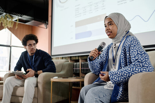 Low angle of young female student wearing hijab sitting in armchair speaking at conference in modern college