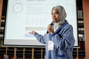 Medium shot of young female university student wearing hijab holding microphone doing presentation,...