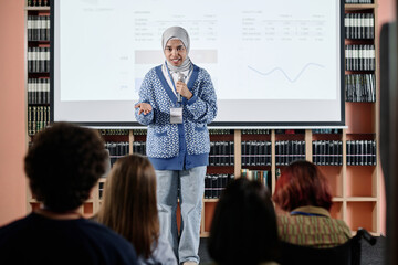 Confident Muslim female student standing in front of audience doing presentation in university library, copy space