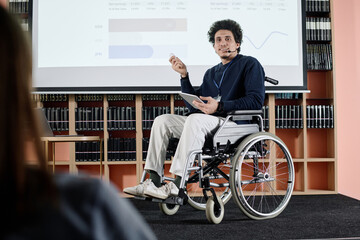 Over-the-shoulder shot of Middle Eastern student in wheelchair holding digital tablet speaking at...