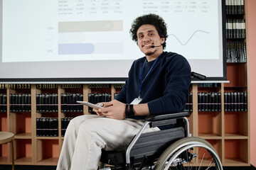 Portrait of cheerful young man with disability in wheelchair holding digital tablet smiling at...