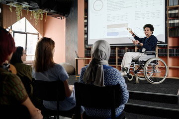 Cheerful Middle Eastern student with disability in wheelchair doing presentation at conference in university library